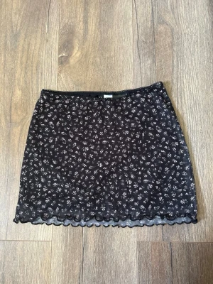 Svart blommig kortkjol med spetskant - Söt svart kortkjol med små vita blommor och fin spetskant längst ner. Kjolen har en rak passform och är perfekt för dig som gillar en feminin och trendig stil. Tunn och lätt, passar bra till både sneakers och boots.