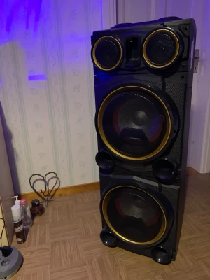 Stor trådlös högtalare med DJ-panel - Säljer en stor trådlös högtalare från Andersson med DJ-panel på ovansidan. Högtalaren är svart med guldfärgade ringar runt elementen och har flera stora högtalarelement för kraftigt ljud. Perfekt för dig som vill ha rejält tryck på musiken och gillar att mixa.