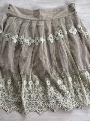 Beige kjol med spets och blommor - Söt beige kjol från EPA Company i storlek L. Kjolen har ett lager av tunn mesh med broderade blommor och spetsdetaljer längs nederkanten. Perfekt för dig som gillar romantisk och feminin stil.