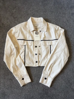 Vit cropped jeansjacka med svarta detaljer - Snygg vit cropped jeansjacka med svarta detaljer och knappar framtill. Perfekt för dig som vill ha en stilren och trendig look.