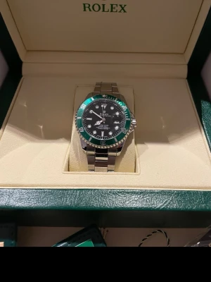 Klocka med grön bezel - Säljer en stilren med svart urtavla och grön bezel. Klockan har ett klassiskt armband i silverfärgat stål och tydliga indexmarkeringar. Perfekt för dig som vill ha en ikonisk och tidlös accessoar.