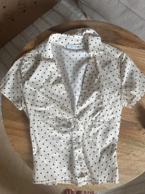 Prickig skjorta design by si - Snygg vit blus med svarta prickar och korta ärmar. Blusen har en klassisk krage, knäppning framtill och markerad midja för en feminin siluett. Perfekt till jeans eller kjol för en trendig look.