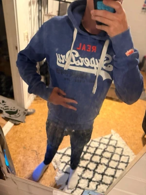 Blå Superdry hoodie med tryck - Säljer en blå hoodie från Superdry med vit och röd logga på bröstet och vita dragsnören i huvan. Hoodien har en klassisk passform, ribbade muddar och en stor ficka framtill. Perfekt för dig som gillar streetwear och vill ha något bekvämt och snyggt.