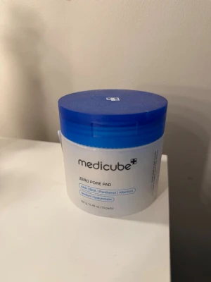 Medicube Zero Pore Pad toner pads - Medicube Zero Pore Pad är runda toner pads i en vit burk med blått lock. Innehåller AHA, BHA, panthenol, allantoin och sodium hyaluronate. Perfekt för att fräscha upp huden och minimera porer. Kommer i en praktisk plastburk.