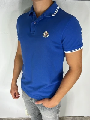 Moncler Piké - Snygg blå pikétröja med vit kant på krage och ärmslut. Tröjan har korta ärmar, knappslå och en broderad logga på bröstet. Perfekt till jeans för en clean och fräsch look.