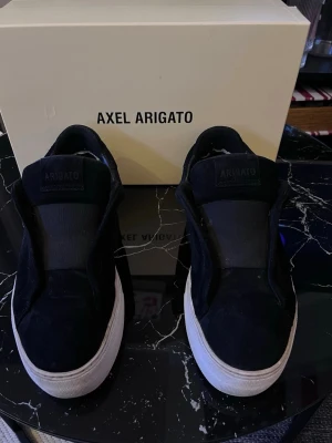 Svarta sneakers från Axel Arigato - Stilrena svarta sneakers från Axel Arigato i mjuk mocka med bred plös och diskret logga på sidan. Skorna har en chunky vit sula och minimalistisk design utan synliga snören. Storlek 41, men passar även dig som vanligtvis har 42. Perfekta för dig som gillar en clean och modern look. Originalbox följer med. Kan tvätta skorna innan jag skickar. Pris kan diskuteras vid snabb affär, hör gärna av er om ni undrar något! 😊