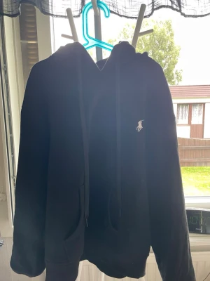 Svart hoodie med Polo Ralph Lauren-logo - Snygg svart hoodie från Polo Ralph Lauren med klassisk vit broderad logga på bröstet. Hoodien har huva med dragsko och en stor magficka framtill. Snörerna går enkelt att ta bort eller stoppa in om man inte gillar dom, hör av er vid minsta fundering eller ifall ni vill ha mer bilder. Pris kan diskuteras