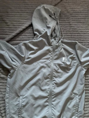 Ljusgrå vindjacka från Under Armour - Snygg ljusgrå vindjacka från Under Armour med dragkedja framtill och huva med dragsko. Jackan har två sidofickor och elastiska muddar vid ärmsluten. Perfekt för blåsiga dagar och sportiga outfits.