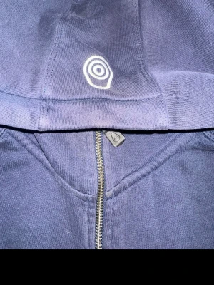 Blå hoodie från Sail Racing - Snygg Blå hoodie från Sail Racing med dragkedja framtill och justerbar huva med vita snören där det står 'Sail Racing'. Broderad logga på huvan och mjukt material som känns skönt mot huden. Perfekt för en avslappnad och cool stil. OBS! Skador finns, längst ner på armen har den gått sönder( går dock att reparera med nål & tråd ) Finns också fläckar som jag inte testat ta bort då jag köpte den second Hand i detta skicket och aldrig fått tiden att försöka fixa den, som var tanken vid köpet. 🌷🤩