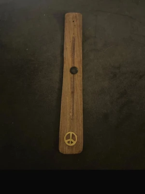 Rökelsehållare i trä med peace-märke - Säljer en brun rökelsehållare i trä med ett inlagt gult peace-märke längst ner. Perfekt för att hålla rökelsepinnar på plats och skapa en avslappnad vibe i rummet. Enkel och stilren design som passar de flesta inredningar.