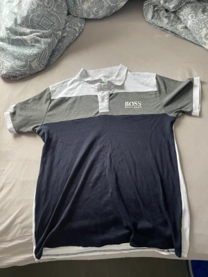 Flerfärgad piké från Hugo Boss - Snygg pikétröja från Hugo Boss i vitt, grått och mörkblått. Klassisk krage med knappar och logga på bröstet. Tillverkad i mjuk bomull som känns skön mot huden. Perfekt för en clean och stilren look.