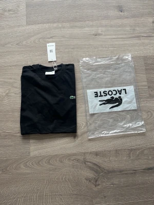 Svart t-shirt från Lacoste - Klassisk svart t-shirt från Lacoste med den ikoniska gröna krokodilloggan broderad på bröstet. T-shirten har rund halsringning och korta ärmar. Tillverkad i mjuk bomull för en skön känsla. Perfekt basplagg till garderoben.