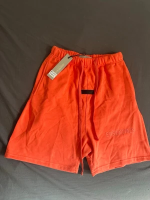 Korall shorts från Essentials - Säljer ett par korall färjade shorts från Essentials med broderad logga på benet. Shortsen har elastisk midja med snörning och är tillverkade i mjuk bomull. Perfekta för sommaren och riktigt sköna att bära.