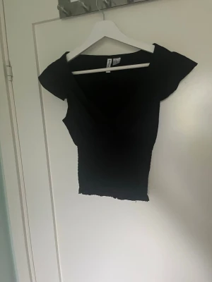 Svart croppad v-ringad topp - Snygg svart croppad topp med v-ringning och korta ärmar. Perfekt att styla med jeans eller kjol för en trendig look. Toppens material känns mjukt och stretchigt, vilket gör den bekväm att bära hela dagen.