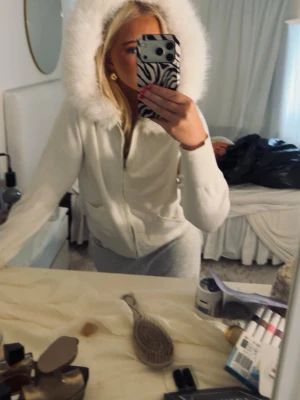 Vit hoodie med fluffig huva - Säljer en vit hoodie med dragkedja och stor fluffig huva med fuskpäls runt kanten. Hoodien har två fickor framtill och är i mjukt material, perfekt för en chill och cozy look.