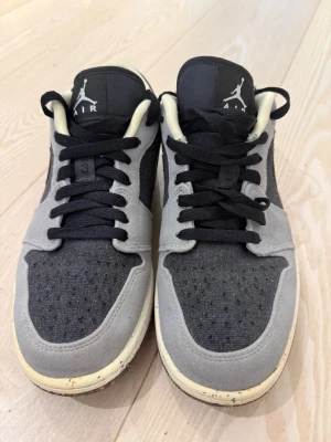 Gråa Air Jordan 1 Low sneakers - Snygga Air Jordan 1 Low sneakers i grått och svart med svart swoosh och blå detaljer på hälen. Ovandelen är i textil och mocka, med svart snörning och vit mellansula med stänk. Perfekta för dig som gillar streetwear och vill ha något unikt.