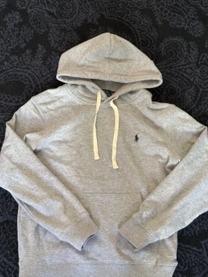 Grå hoodie från Polo  - Snygg grå hoodie från Polo Ralph Lauren med klassisk huva och vita dragsnören. Tröjan har en broderad logga på bröstet och en stor magficka. Tillverkad i mjukt bomullsmaterial som är perfekt för chill dagar. Enkel och stilren design som funkar till det mesta.