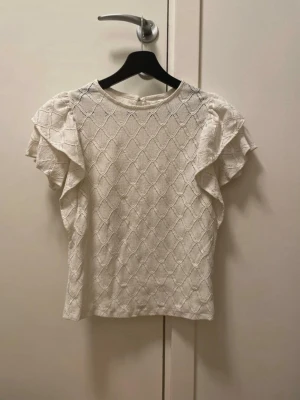 Vit blus med volangärmar - Supersöt vit blus med volangdetaljer på ärmarna och ett diskret mönster över hela tyget. Blusen har rund halsringning och är kortärmad, perfekt för en fräsch och stilren look. Passar till både jeans och kjol för en trendig outfit.