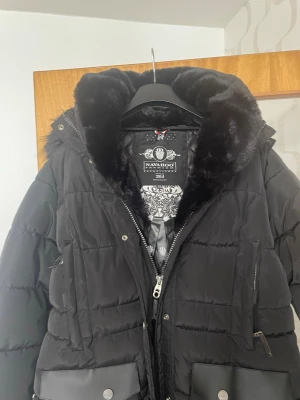 Svart lång dunjacka med fuskpäls - Säljer en svart lång dunjacka från Navahoo med stor huva fodrad i mjuk fuskpäls. Jackan har dragkedja, tryckknappar, flera fickor med dragkedja och lock samt detaljer i skinnimitation. Perfekt för kalla vinterdagar och riktigt snygg design.