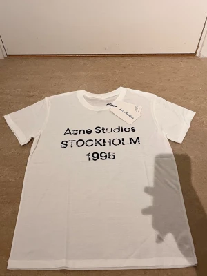 Vit Acne Studios t-shirt Stockholm - Vit t-shirt från Acne Studios med svart text 'Acne Studios STOCKHOLM  1996' på bröstet. Klassisk rund hals och korta ärmar. Tillverkad i mjuk bomull för en clean och stilren look.