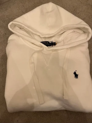 Vit hoodie från Polo Ralph Lauren - Säljer en stilren vit hoodie från Polo Ralph Lauren med klassisk huva och dragsnören. Tröjan har det ikoniska broderade logotypen i svart på bröstet och ribbade muddar vid ärmar och nederkant. Perfekt för en avslappnad och trendig look.Priset går att diskutera vid snabb affär, finn de frågor så är de bara o skriv 😊