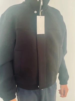 Svart bomberjacka från Zara - Snygg svart bomberjacka från Zara med hög krage, dragkedja och tryckknappar framtill. Jackan har två sidofickor och ribbade muddar vid ärmslut och nederkant. Stilren och enkel design som passar till många olika outfits.