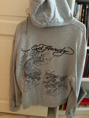 Grå Ed Hardy hoodie med drakmotiv - Snygg grå zip-hoodie från Ed Hardy med stora drakmotiv i svart på både fram- och baksida. Hoodien har huva med dragsko, två fickor fram och coola detaljer på ärmarna. Perfekt för dig som gillar streetwear och vill sticka ut lite extra.
