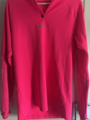 Rosa långärmad träningströja Nike - Snygg rosa långärmad träningströja från Nike med halv dragkedja framtill och reflekterande logga på bröstet. Tröjan har diskret mönster på ärmarna och är tillverkad i ett lätt, stretchigt material som passar perfekt för löpning eller gym.