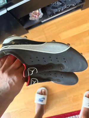 Svarta fotbollsskor med vita detaljer - Säljer ett par svarta fotbollsskor från Puma med vita och röda detaljer. Skorna har klassisk snörning, dobbar för gräsplan och ventilerande hål på sidorna. Perfekta för dig som vill ha stilrena och funktionella skor på planen.