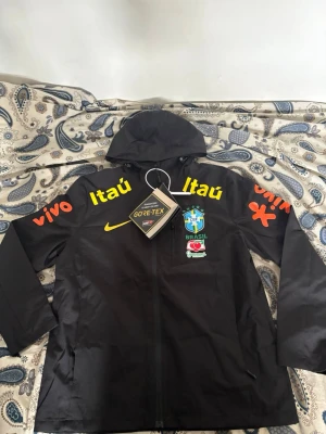 Svart Nike Brasil vindjacka med luva HELT NY! - Snygg svart vindjacka från Nike med Brasil-logga och sponsormärken i gult och orange. Jackan har huva, dragkedja framtill och är tillverkad i GORE-TEX-material som andas. Perfekt för dig som gillar sportig stil och vill sticka ut. Helt ny