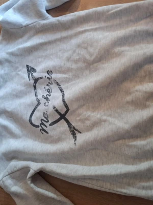 Ljusgrå hoodie med tryck från Gina Tricot - Ljusgrå hoodie från Gina Tricot med svart tryck på ryggen som föreställer ett hjärta med pil och texten 'ma chérie'. Mjuk insida i fleece och klassisk huva. Perfekt för en chill och avslappnad stil.