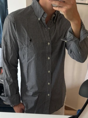 Svartvit rutig skjorta med knappar - Klassisk svartvit rutig skjorta med knappar framtill och button-down krage. Skjortan har lång ärm och en liten broderad logga på bröstet. Tillverkad i bomull för en bekväm känsla och passar perfekt till jeans eller chinos.