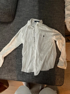 Vit Ralph lauren skjorta - Klassisk vit skjorta med lång ärm och knappar framtill. Skjortan har en krage och en liten broderad logga på bröstet. Perfekt till många olika stilar och enkel att matcha med andra plagg.