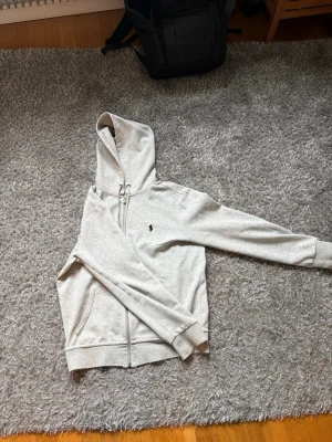 Grå hoodie från Polo Ralph Lauren - Jag säljer en grå hoodie från Polo Ralph Lauren med dragkedja och huva för endast 299 kr. Tröjan är i storlek S och har igensydda hål bak på tröjan som ni ser på bilden, annars ett mycket bra skick, hör av er vid funderingar!