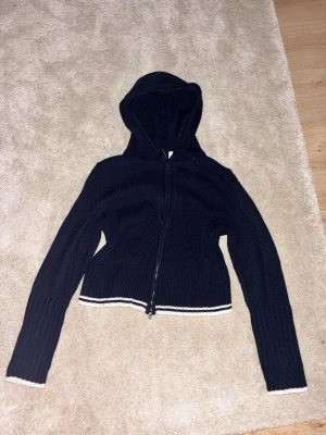 Svart stickad hoodie med dragkedja - Snygg svart stickad hoodie med dragkedja framtill och vita ränder längst ner på ärmar och midja. Tröjan har en klassisk huva och ribbade detaljer som ger en cool och avslappnad look. Perfekt till jeans eller kjol för en trendig vardagsstil.