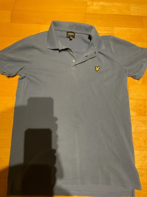 Blå pikétröja från Lyle & Scott - Klassisk blå pikétröja från Lyle & Scott Junior med gul logga på bröstet. Tröjan har krage, korta ärmar och knappar vid halsen. Perfekt för en avslappnad och stilren look.