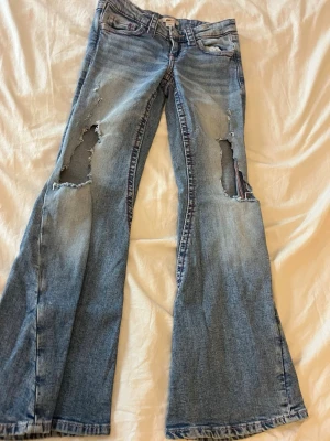 Blå slitna bootcut jeans med hål - Ett par ljusblå bootcut jeans med stora slitningar och hål på båda benen. Jeansen har klassisk dekorativ bakficka med broderi. Sällan använda!