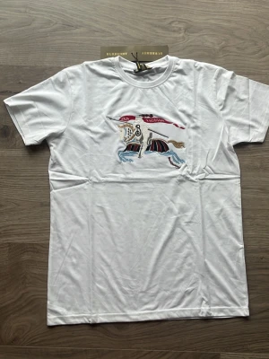 Vit Burberry t-shirt med broderi - Säljer en vit t-shirt från Burberry med ett färgglatt broderat motiv på bröstet. T-shirten är i 100% bomull och har klassisk passform med korta ärmar och rund hals. Perfekt för dig som vill ha något stilrent men ändå unikt.