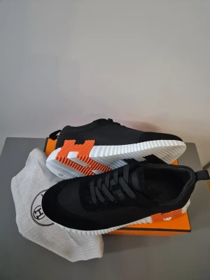 H skor - Snygga svarta sneakers med ovandel i textil och detaljer i mocka. Sulan är vit med orange och svarta inslag som ger en cool kontrast. Skorna har klassisk snörning och låg profil. Perfekta för dig som gillar en sportig och modern stil.