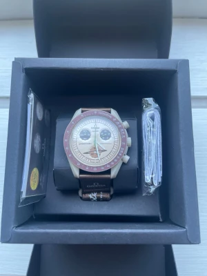Omega x Swatch Saturn - Snygg klocka från Omega x Swatch med beige urtavla, bruna detaljer och brunt armband. Klockan har kronograf-funktion och tydliga markeringar runt urtavlan. Kommer i originalbox med tillbehör och manual.