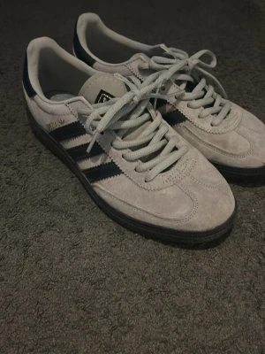 Adidas Gazelle grå sneakers - Grå Adidas Gazelle sneakers i mocka med svarta klassiska ränder på sidorna och svart sula. Skorna har snörning och gulddetaljer på sidan och plösen. Storleken stämmer inte in med vad det står i skon som är UK 5 men den riktiga storleken ska vara UK 5,5! Mått i cm på sista bilden och det finns lite fläckar på skorna och kom dm för mer information och priset går även att diskuteras vid snabb affär!!