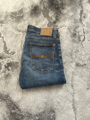 Blå jeans från Nudie Jeans - Säljer ett par klassiska blå jeans från Nudie Jeans med snygga slitningar och kontrastsömmar. Modellen har raka ben och råa kanter vid bensluten. Jeansen har fem fickor och är tillverkade i denim med en cool vintagekänsla.