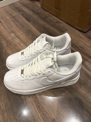  Nike Air Force 1 sneakers storlek 43 - Klassiska vita Nike Air Force 1 sneakers i lågt utförande. Skorna har en stilren design med perforerad tå, vit sula och ikoniska Swoosh-loggan på sidan. Tillverkade i slittåligt läder med vita skosnören och metallisk detalj framtill.