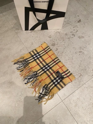 Gul/beige rutig halsduk från Burberry - Klassisk gul/beige halsduk från Burberry med ikoniskt rutmönster i svart, vitt och rött. Halsduken har tvinnade fransar i ändarna och är tillverkad i mjuk ull. Perfekt accessoar för att lyfta din outfit med en tidlös och lyxig känsla.
