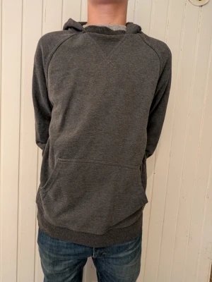 Mörkgrå hoodie med magficka från Tiger of sweden - En enkel och stilren mörkgrå hoodie med huva och stor magficka framtill. Tillverkad i mjukt sweatshirtmaterial och har ribbade muddar vid ärmslut och nederkant. Liten tygpatch med logga vid fickan för en diskret detalj.
