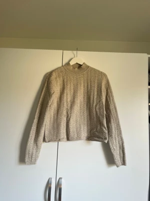 Beige kabelstickad tröja från Lindex - Säljer en beige kabelstickad tröja från Lindex med rund hals och långa ärmar. Tröjan har ett klassiskt kabelmönster och ribbade muddar vid hals, ärmslut och nederkant. Perfekt för lager på lager och enkel att matcha med jeans eller kjol.