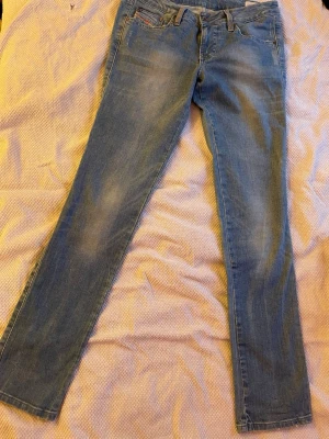 Ljusblå raka jeans från Diesel - Säljer ett par ljusblå jeans från Diesel med raka ben och klassisk femficksmodell. Jeansen har snygga slitningar och detaljerade sömmar samt metallknappar och nitar. Perfekta för en avslappnad och trendig look.