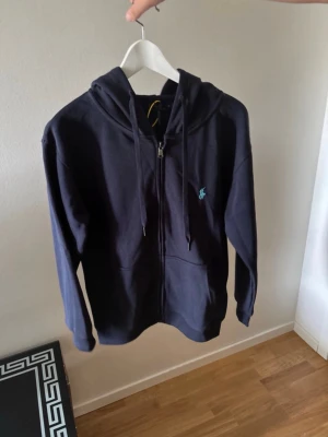 Larph lauren - Mörkblå hoodie med dragkedja och huva. Klassisk design med fickor framtill och justerbara snören i huvan. Liten broderad logga i ljusblått på bröstet. Tillverkad i mjukt sweatshirtmaterial, perfekt för chill dagar.
