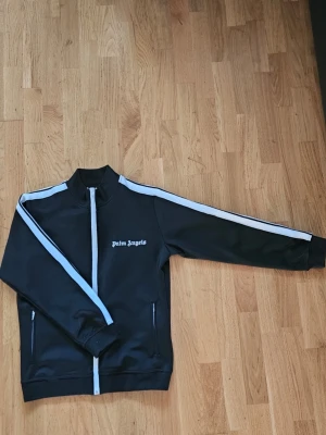 Svart track jacket från Palm Angels - Snygg svart track jacket från Palm Angels med vita ränder längs ärmarna och dragkedja framtill. Jackan har ståkrage, ribbade muddar och två fickor med dragkedja. Perfekt för en sportig och trendig look.
