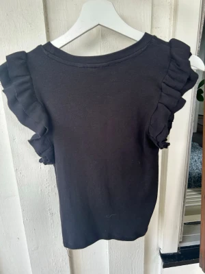 Svart ärmlös topp med volang - Snygg svart topp i ribbad bomull med volangdetaljer längs axlarna. Modellen är ärmlös och har en rund halsringning. Perfekt att styla med jeans eller kjol för en trendig look.